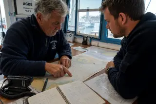 Two generations of local boaters in a Mediterranean capitainerie, reviewing checklists and nautical charts by natural light, focused on preparation and shared expertise.
