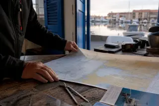 Weathered hands of a mariner spreading a marine weather chart in a traditional Mediterranean harbor office at dawn, with navigation tools and subtle port view through blue shutters.