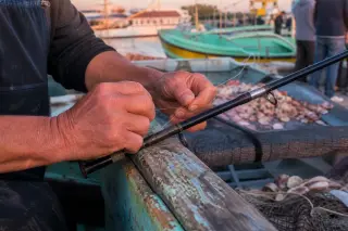 Two experienced fishers' hands arranging jigs and charts on a small boat's wooden table in the early morning, soft Mediterranean light entering through a porthole.