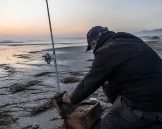 Solitary fisherman at dawn on a windswept Hérault beach, kneeling as he prepares natural bait for surf casting, with soft morning light and authentic shoreline details.