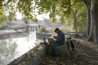 Local fisherman inspecting antique fishing lures by a misty canal lock, with a bicycle and old stonework under Mediterranean morning light.
