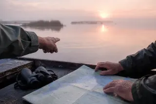 Hands of naturalists pointing at ripples on Thau lagoon at dawn, soft mist, boat edge, map and binoculars, subtle pink morning light.