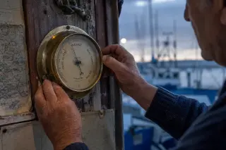 Fisherman's salt-marked hands adjusting a brass barometer on a weathered harbor wall at Cap d'Agde at dawn, with fishing boats and nautical charts in soft Mediterranean morning light.