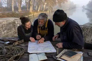 Fishermen and a woman consult marine maps and permits on a canal lock gate in the morning mist, surrounded by fishing gear and paperwork, with soft Mediterranean light and plane trees reflected in still water.