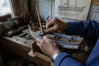 Weathered hands of an older boatbuilder repairing a wooden barge model in a sunlit, cluttered Sète workshop, vintage tools and sketches in the background.
